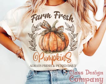 May include: A cream-colored t-shirt featuring a graphic of an orange pumpkin with a leopard print bow, flanked by leafy branches. The text reads Farm Fresh Pumpkins ALWAYS FRESH & PICKED DAILY in brown and orange lettering.