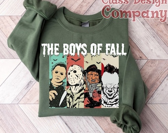 Può includere: Una felpa girocollo verde oliva con la scritta THE BOYS OF FALL in lettere bianche gocciolanti. Sotto il testo ci sono quattro pannelli verticali che raffigurano personaggi di film horror: Michael Myers, Jason Voorhees, Freddy Krueger e Pennywise.