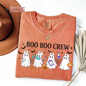 May include: An orange Comfort Colors t-shirt with the text "BOO BOO CREW" and a graphic of cartoon ghosts dressed as medical professionals. The ghosts are wearing nurse hats, holding medical supplies, and surrounded by bats and spiderwebs.