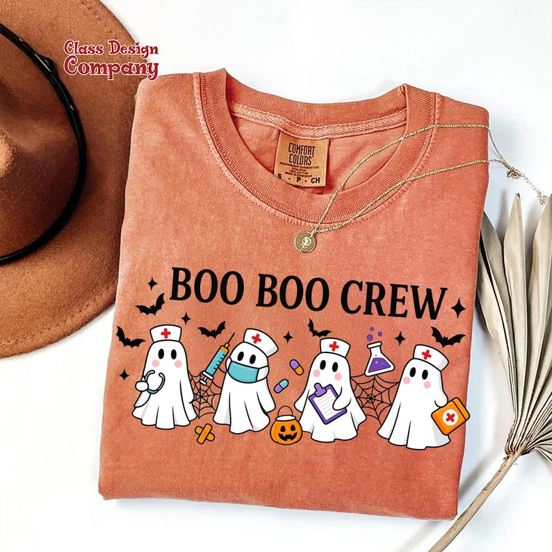 May include: An orange Comfort Colors t-shirt with the text "BOO BOO CREW" and a graphic of cartoon ghosts dressed as medical professionals. The ghosts are wearing nurse hats, holding medical supplies, and surrounded by bats and spiderwebs.