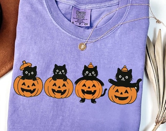May include: A lavender short-sleeve t-shirt featuring a graphic of four black cats sitting in or on orange jack-o'-lantern pumpkins. The shirt has a Comfort Colors label at the neck and the text Glass Design Company in the top left corner.