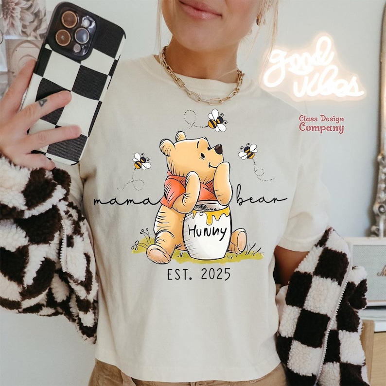 May include: A beige t-shirt with a cartoon Winnie the Pooh illustration holding a honey pot that says "Hunny". The text "mama bear" is above the illustration and "EST. 2025" is below. There are bees flying around Winnie the Pooh.