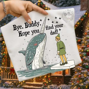 Pode incluir: Camiseta branca com uma narval e um elfo de desenho animado. A narval é cinza e azul com um chifre. O elfo está usando uma roupa verde. O texto na camiseta diz "Bye, Buddy. Hope you find your dad!" Design temático de Natal.