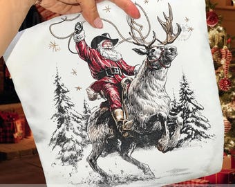 Pode incluir: Um artigo de tecido branco com uma ilustração detalhada em tinta preta e vermelha do Pai Natal com um chapéu de cowboy montado numa rena. Ele segura um laço acima da cabeça, rodeado por pinheiros e pequenas estrelas.