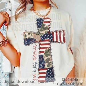 May include: Cream-colored t-shirt featuring a cross design made of patchwork fabric with American flag and camouflage patterns. The words "god bless america" are written in red script. The design is distressed, with frayed edges.