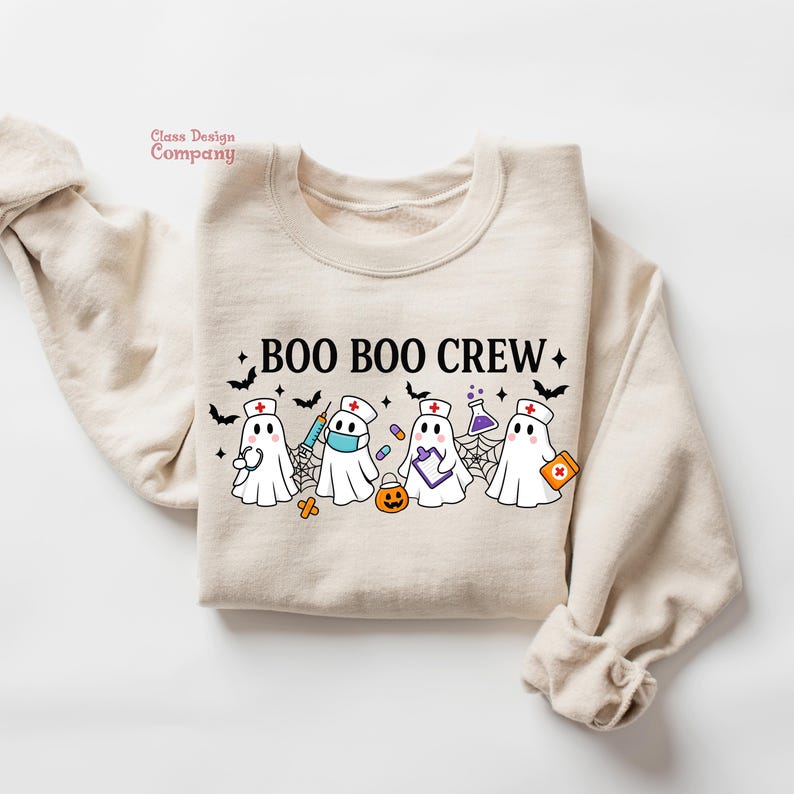 May include: Beige sweatshirt with the text "BOO BOO CREW" and illustrations of ghost characters dressed as nurses, with medical equipment and Halloween elements. The design includes bats, spiderwebs, and a pumpkin.