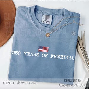 May include: A blue t-shirt with the text "250 YEARS OF FREEDOM" and an American flag graphic. The shirt has a small tag that says "COMFORT COLORS". A gold necklace with a pendant is on the shirt.