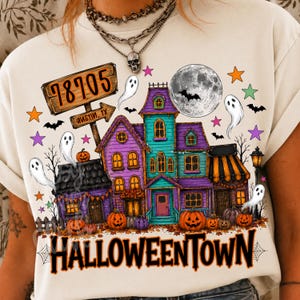 May include: A cream-colored t-shirt featuring a graphic of a haunted house with purple and teal walls, surrounded by ghosts, bats, pumpkins, and stars. A wooden sign reads 78705 Austin, TX. The text HALLOWEENTOWN is printed in large black letters.