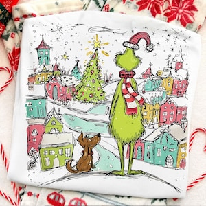 May include: A white t-shirt featuring a colorful graphic illustration of a green creature wearing a red and white striped scarf and hat, standing with a brown dog in front of a decorated Christmas tree and a village of small, multi-colored houses.