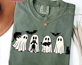 Puede incluir: Una camiseta de manga corta color verde oliva con un estampado gráfico de cuatro fantasmas blancos y gatos negros. El diseño incluye pequeños murciélagos negros y formas de estrellas. La camiseta tiene una etiqueta de marca Comfort Colors.