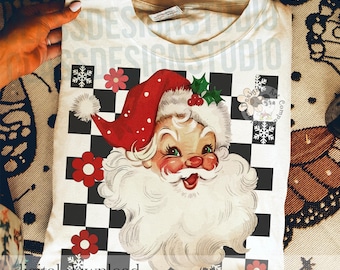 May include: A white t-shirt featuring a graphic of a vintage-style Santa Claus head with a red hat, white beard, and green holly. The background design consists of a black and white checkerboard pattern accented with red flowers and white snowflakes.