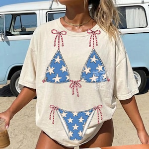 May include: Cream-colored t-shirt featuring a blue bikini design with white stars and red bow accents. The bikini top and bottom are printed on the front of the shirt. The shirt is a casual, relaxed fit.