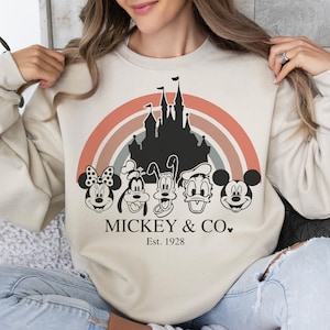 K&ouml;nnte beinhalten: Ein beiger Pullover mit einem Vintage-inspirierten Design mit Mickey Mouse, Minnie Mouse, Donald Duck, Goofy und Pluto. Das Design beinhaltet eine Silhouette von Schloss Aschenputtel und einen Regenbogenbogen. Der Text "MICKEY & CO. Est. 1928" ist unter den Figuren gedruckt.