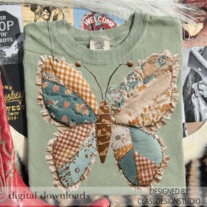 以下が含まれることがあります： セージグリーンのTシャツ。大きな蝶のアップリケが特徴。蝶は茶色、青、クリーム色の様々な生地のパッチでできており、フリルエッジが付いています。「DESIGNED BY CLASSDESIGNSTUDIO」の文字が見えます。