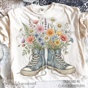 Puede incluir: Camiseta color crema con un diseño floral y zapatillas azules. El gráfico muestra flores coloridas que brotan de un par de zapatillas altas azules. El texto en la camiseta dice "descarga digital" y "diseñado por classdesignstudio."
