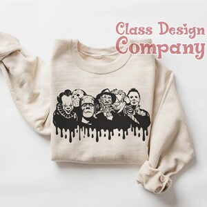 May include: Beige sweatshirt featuring a black graphic of iconic horror movie characters with a dripping effect. The text "Class Design Company" is in pink script. The sweatshirt has long sleeves and a crew neck.