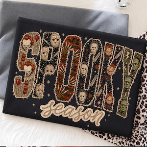 May include: A black t-shirt featuring the word SPOOKY in large, stitched-style block letters filled with horror movie character graphics, including clowns, masked figures, and monsters. Below, the word season is written in a cursive font.