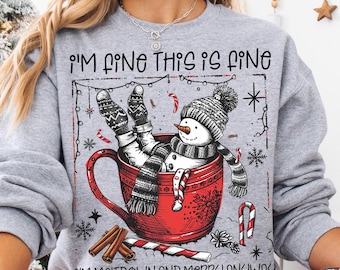 May include: A heather gray sweatshirt featuring a graphic of a snowman sitting in a large red mug. The text reads I'M FINE THIS IS FINE and I'M MELTDOWN AND MERRY ANYWAY. The design includes candy canes, cinnamon sticks, and snowflakes.