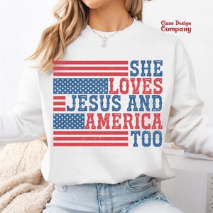 May include: White sweatshirt with a red, white and blue American flag design. The text "She Loves Jesus and America Too" is printed on the sweatshirt.