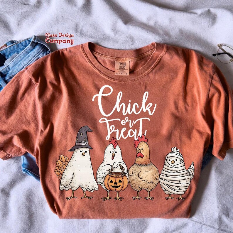 May include: Terracotta-colored t-shirt with the text "Chick or Treat" and a graphic of four chickens in Halloween costumes. One chicken wears a witch hat, another holds a pumpkin bucket, and a third is wrapped in bandages.
