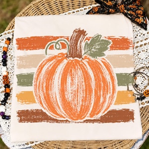 May include: A cream-coloured t-shirt featuring a large, textured orange pumpkin graphic centred over horizontal brushstrokes in shades of orange, tan, olive green, and brown. The pumpkin has a brown stem and a single green leaf.