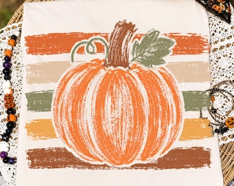 Può includere: Una t-shirt color crema con una grande grafica di una zucca arancione testurizzata, centrata sopra pennellate orizzontali nei toni dell'arancione, beige, verde oliva e marrone. La zucca ha un picciolo marrone e una foglia verde.