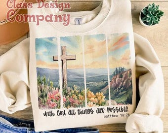May include: A cream-coloured crewneck sweatshirt featuring a watercolour-style graphic of a cross in a landscape with flowers and mountains. The text reads, "With God all things are possible" and "Matthew 19:26" in a dark script font.