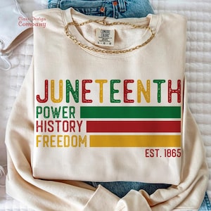 Peut inclure: Un sweat-shirt crème avec les mots "JUNETEENTH", "POWER", "HISTORY", "FREEDOM" et "EST. 1865" imprimés en rouge, jaune, vert et noir.