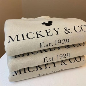 K&ouml;nnte beinhalten: Ein Stapel aus drei beigen Sweatshirts mit schwarzem Text, der "Mickey & Co. Est. 1928" lautet. Das oberste Sweatshirt hat eine schwarze Mickey-Maus-Silhouette.