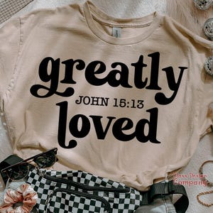 May include: A beige t-shirt with the text "greatly loved" in black, retro-style font. The text "JOHN 15:13" is printed in smaller black font below the main text.