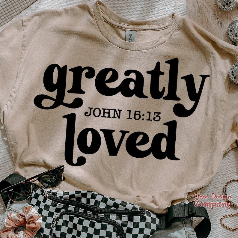 May include: A beige t-shirt with the text "greatly loved" in black, retro-style font. The text "JOHN 15:13" is printed in smaller black font below the main text.