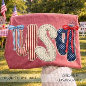 Puede incluir: Una camiseta roja con la palabra "USA" en un dise&ntilde;o patri&oacute;tico. La "U" tiene un lazo azul, la "S" es de punto de cable y la "A" tiene un lazo rojo. La camiseta es una descarga digital.