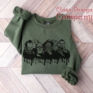 May include: Olive green sweatshirt featuring a black graphic of horror movie characters with a dripping effect. The characters include Pennywise, Jason Voorhees, Frankenstein's monster, Freddy Krueger, and Michael Myers. The text "Class Design Company" is in the upper right corner.