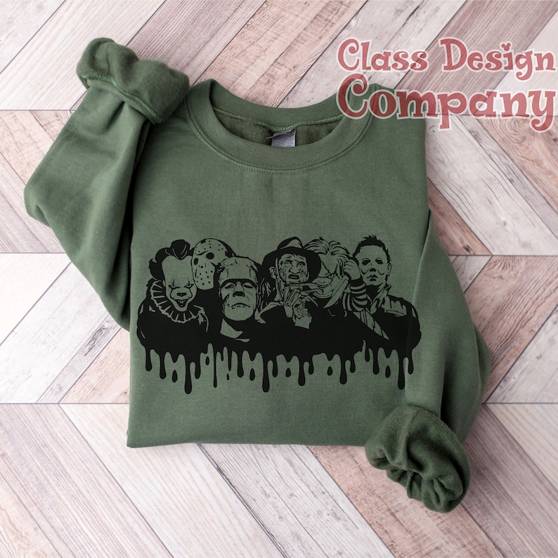 May include: Olive green sweatshirt featuring a black graphic of horror movie characters with a dripping effect. The characters include Pennywise, Jason Voorhees, Frankenstein's monster, Freddy Krueger, and Michael Myers. The text "Class Design Company" is in the upper right corner.