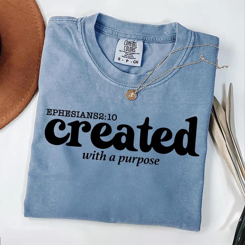 May include: A light blue t-shirt with the text "EPHESIANS 2:10 created with a purpose" printed in black. The shirt has a distressed look and is made of a soft, comfortable material.