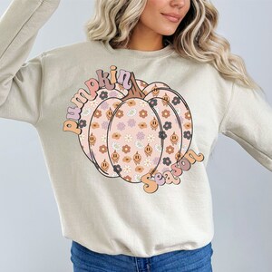 May include: Beige sweatshirt with a pumpkin graphic, decorated with flowers and smiley faces. The words "pumpkin season" are arched above and below the pumpkin in a retro font. A cosy autumn-themed garment.