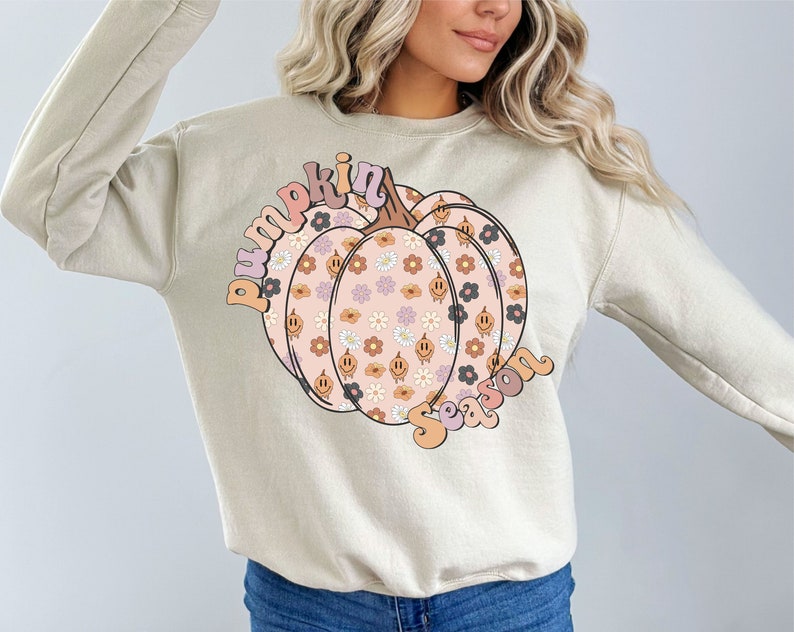 May include: Beige sweatshirt with a pumpkin graphic, decorated with flowers and smiley faces. The words "pumpkin season" are arched above and below the pumpkin in a retro font. A cosy autumn-themed garment.