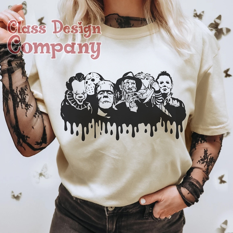 May include: Cream-colored t-shirt featuring a black graphic of iconic horror movie characters, including Pennywise, Jason Voorhees, and Michael Myers. The graphic has a dripping effect at the bottom. The text "Class Design Company" is in the upper left corner.