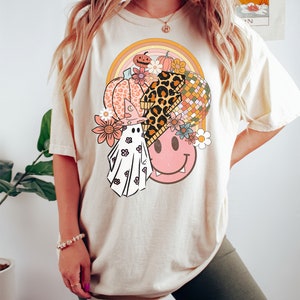 May include: Cream-coloured t-shirt with a vibrant graphic design. The design incorporates a pumpkin, flowers, a disco ball, a ghost, and a smiley face. The t-shirt has a relaxed fit and short sleeves.