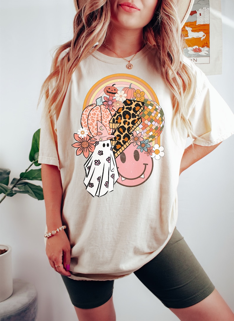 May include: Cream-coloured t-shirt with a vibrant graphic design. The design incorporates a pumpkin, flowers, a disco ball, a ghost, and a smiley face. The t-shirt has a relaxed fit and short sleeves.