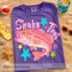 Pode incluir: Camiseta roxa com um gráfico de peixe e as palavras "Shake That" em rosa. O peixe é laranja e rosa com estrelas azuis, amarelas e rosas. A camiseta está em uma superfície arenosa com acessórios de praia.