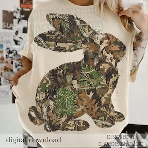 Op de afbeelding: Crèmekleurig T-shirt met een grote illustratie van een konijn in camouflagepatroon. Het konijnontwerp gebruikt bruin-, groen- en beigetinten, met blad- en takdetails. De woorden "digital download" staan linksonder.