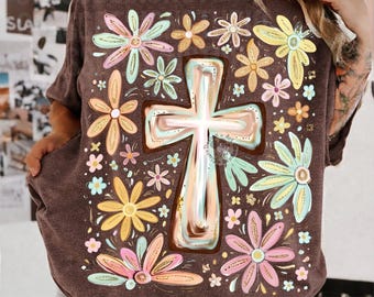 May include: A brown short-sleeve t-shirt featuring a large central cross graphic with a metallic-look finish, surrounded by a pattern of stylized flowers in shades of pink, yellow, light blue, and orange across the front.