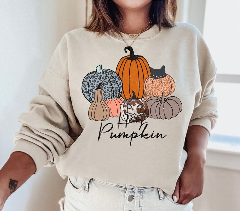 May include: Beige sweatshirt with a graphic of pumpkins in orange, leopard print, and blue leopard print. The text "Hello Pumpkin" is written in black script below the pumpkins. A seasonal autumn design.