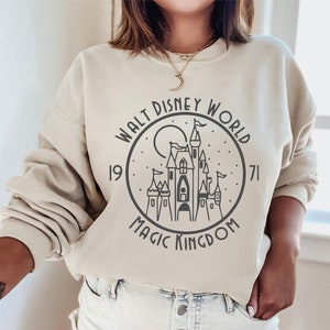 K&ouml;nnte beinhalten: Ein beiger Pullover mit einem schwarz-wei&szlig;en Grafikdruck von Cinderellas Schloss. Die Grafik enth&auml;lt den Text "Walt Disney World 1971 Magic Kingdom".