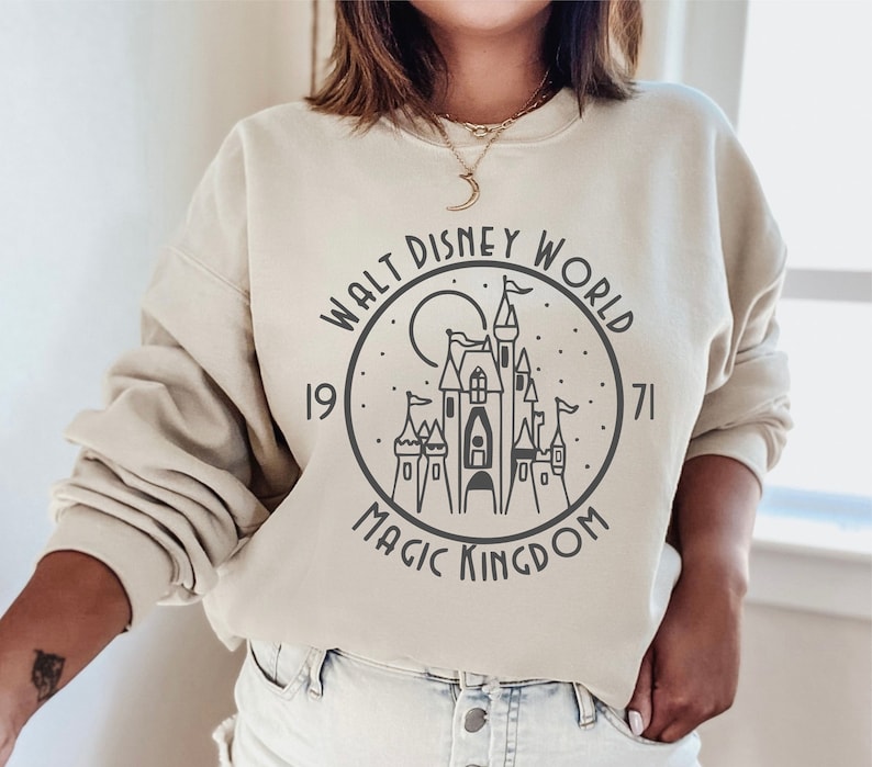 K&ouml;nnte beinhalten: Ein beiger Pullover mit einem schwarz-wei&szlig;en Grafikdruck von Cinderellas Schloss. Die Grafik enth&auml;lt den Text "Walt Disney World 1971 Magic Kingdom".