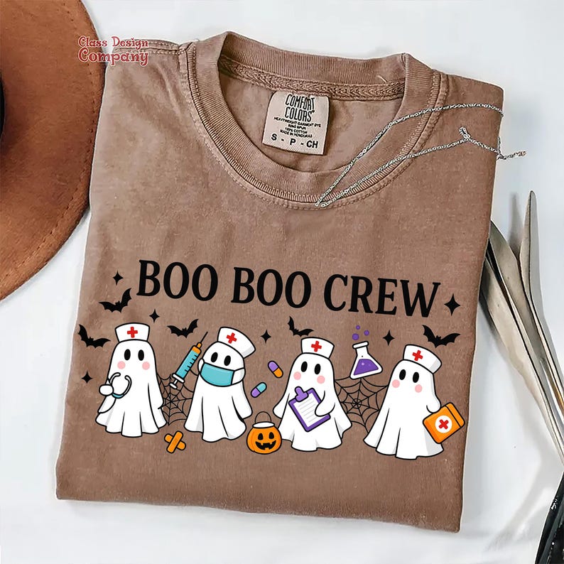 May include: A brown Comfort Colors t-shirt with the text "BOO BOO CREW" and illustrations of ghost characters dressed as nurses, with bats and spiderwebs. The ghosts are holding medical tools and supplies.