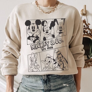 K&ouml;nnte beinhalten: Ein beiger Pullover mit einem schwarz-wei&szlig;en Grafikdruck von Mickey Mouse, Minnie Mouse, Donald Duck und Goofy. Die Grafik enth&auml;lt den Text "Mickey & Co. Est. 1928".