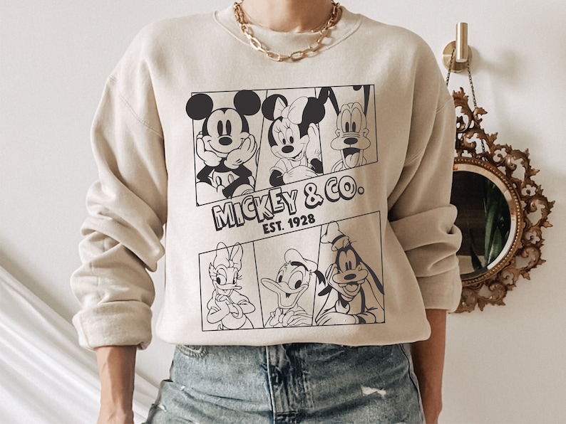 K&ouml;nnte beinhalten: Ein beiger Pullover mit einem schwarz-wei&szlig;en Grafikdruck von Mickey Mouse, Minnie Mouse, Donald Duck und Goofy. Die Grafik enth&auml;lt den Text "Mickey & Co. Est. 1928".