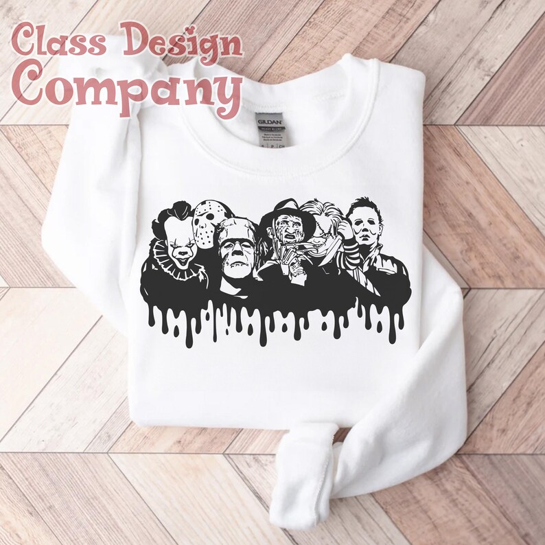May include: White sweatshirt featuring a black graphic of iconic horror movie characters with a dripping effect. The text "Class Design Company" is in the upper left corner. The sweatshirt is folded on a wooden surface.